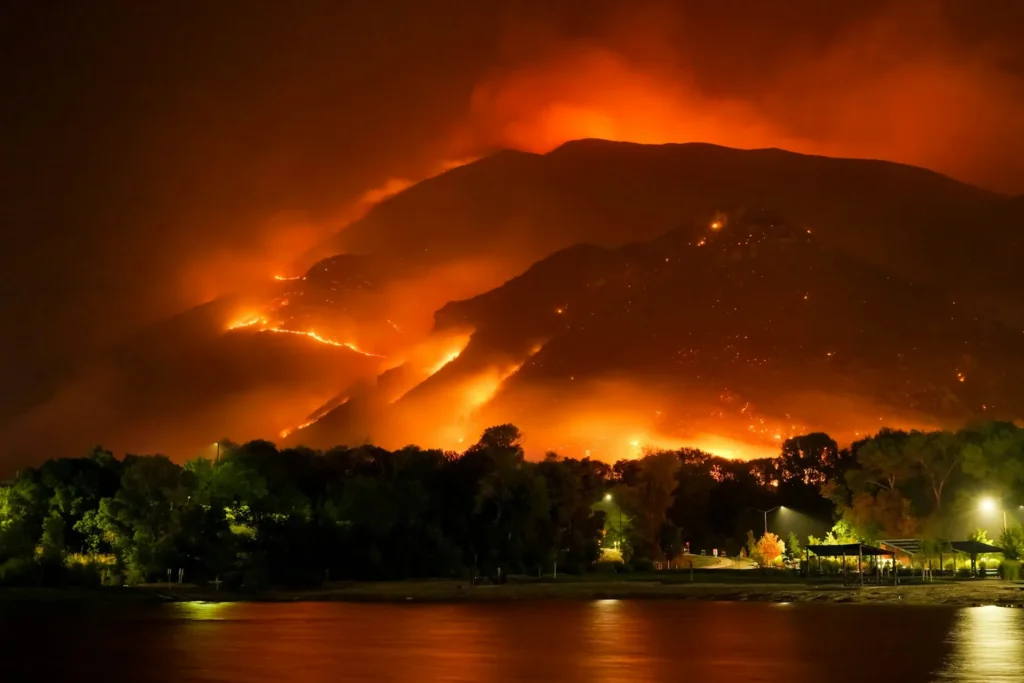 A massive wildfire rages across a mountain ridge at night, illuminating the sky with an intense orange glow and dense smoke.